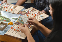 Load image into Gallery viewer, Young woman player holding two playing cards above her player tracker while playing the board game Bharata 600 BC